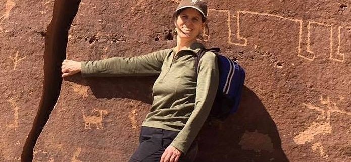 Liesl Dees, UUCB Ministerial Intern, white woman smiling with brown hair, standing with her hand in the crack of a rock that has Native American drawings of animals