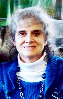 White woman with light gray hair, deep beautiful eyes, smiling, wearing a blue sweater jacket