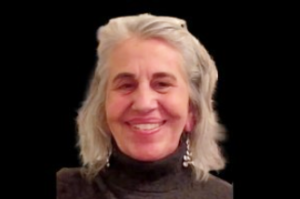 White woman with light gray hair and big smile with a brown turtleneck sweater shirt