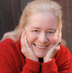 a Caucasian woman with strawberry and gray gold hair wearing a red sweater, holding her hands to her face and smiling.